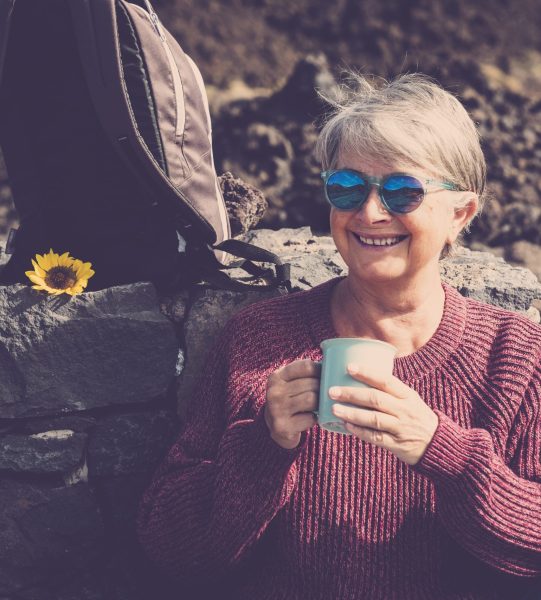 Cheerful happy caucasian aged mature woman enjoy a hot beverage like tea or coffee resting after a trekking day of outdoor leisure activity at the mountain - alternative vacation lifestyle